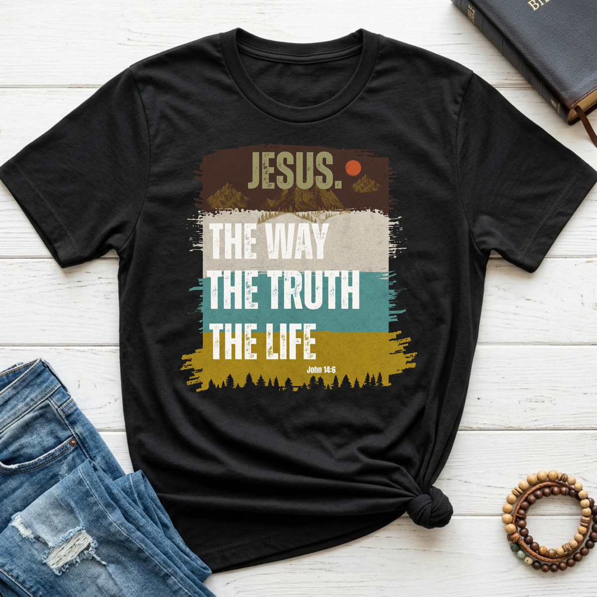 A black graphic t-shirt laid flat on a white wood background, styled with blue jeans, a book, and a beaded bracelet. The t-shirt features a retro-style layered graphic with the text 'JESUS.