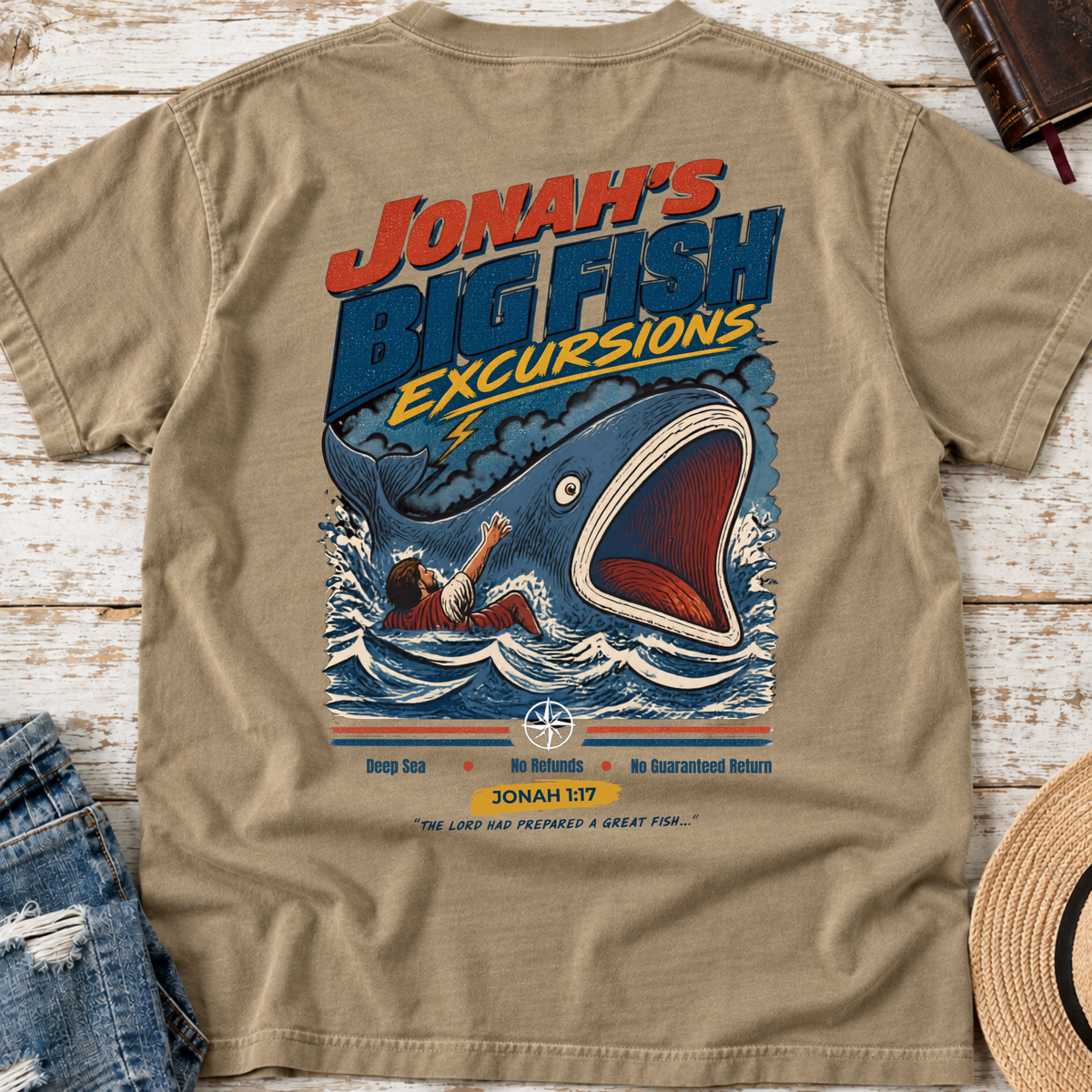 A khaki christian graphic t-shirt laid flat on a white wood background, showing the back side with a colorful illustration of a giant fish and the text 'Jonah's Big Fish Excursions'.