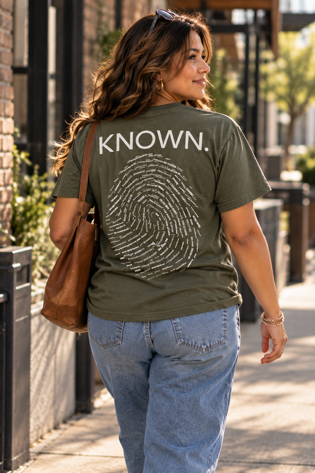A woman wearing a military green Christian graphic t-shirt by SeedSowerGraphics with 'KNOWN.' and a fingerprint graphic printed on the back, walking outdoors on a sidewalk.