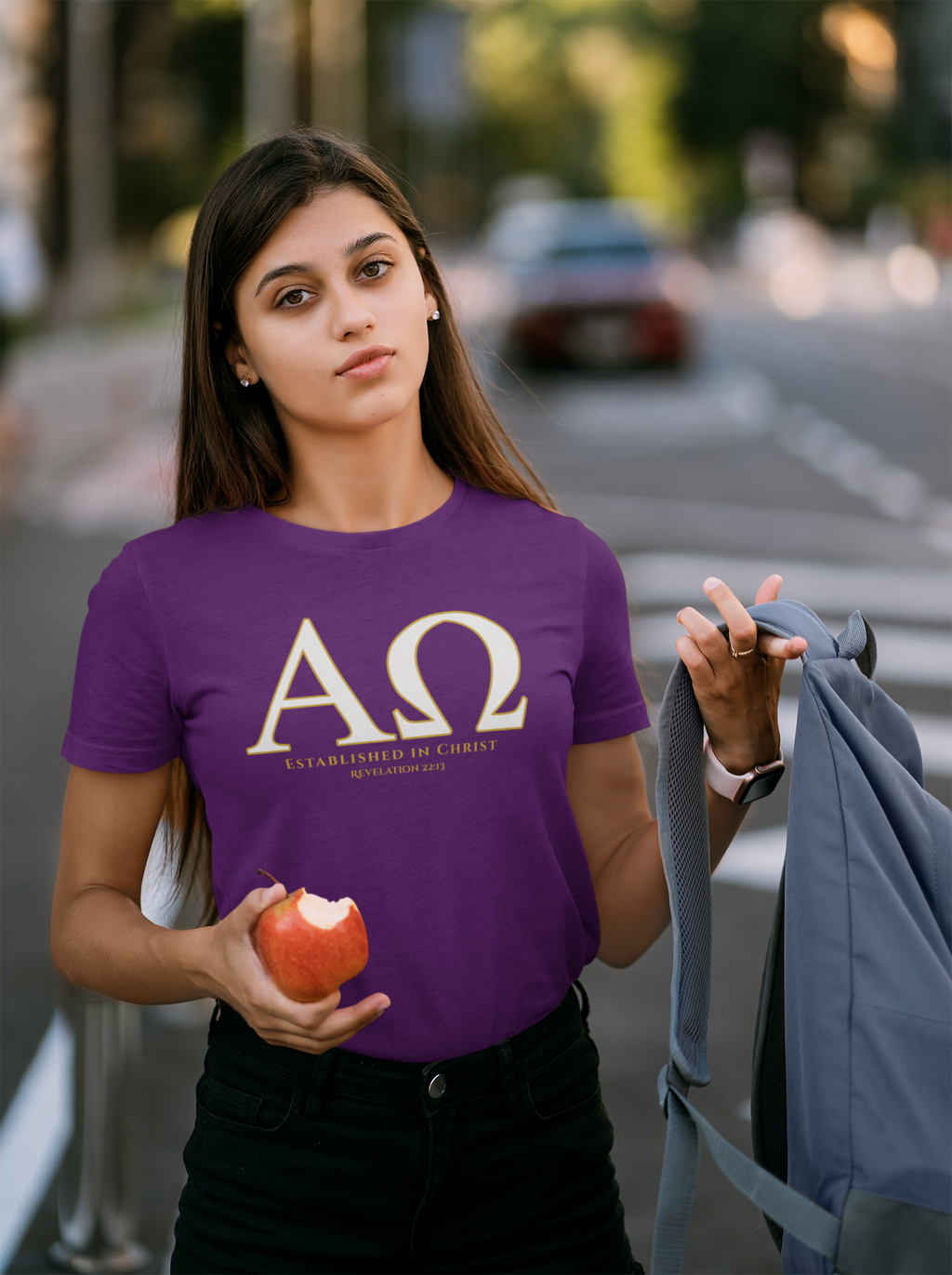 A woman wearing a purple t-shirt with a large gold Greek letter 'ΑΩ' (Alpha Omega) and the motto 'ESTABLISED IN CHRIST' printed on the front. She is holding a red apple and a gray backpack, standing outdoors on a blurred street background.