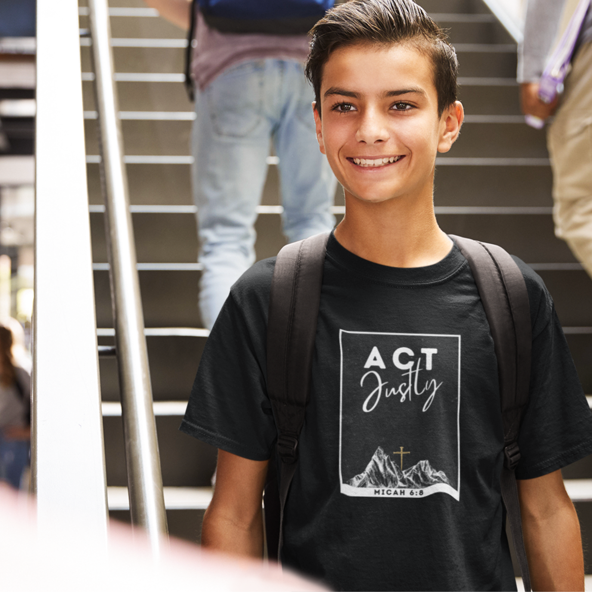 A smiling young man wearing a black graphic t-shirt with 'ACT Justly MICAH 8:5' printed on the front, standing on outdoor stairs with a backpack on.
