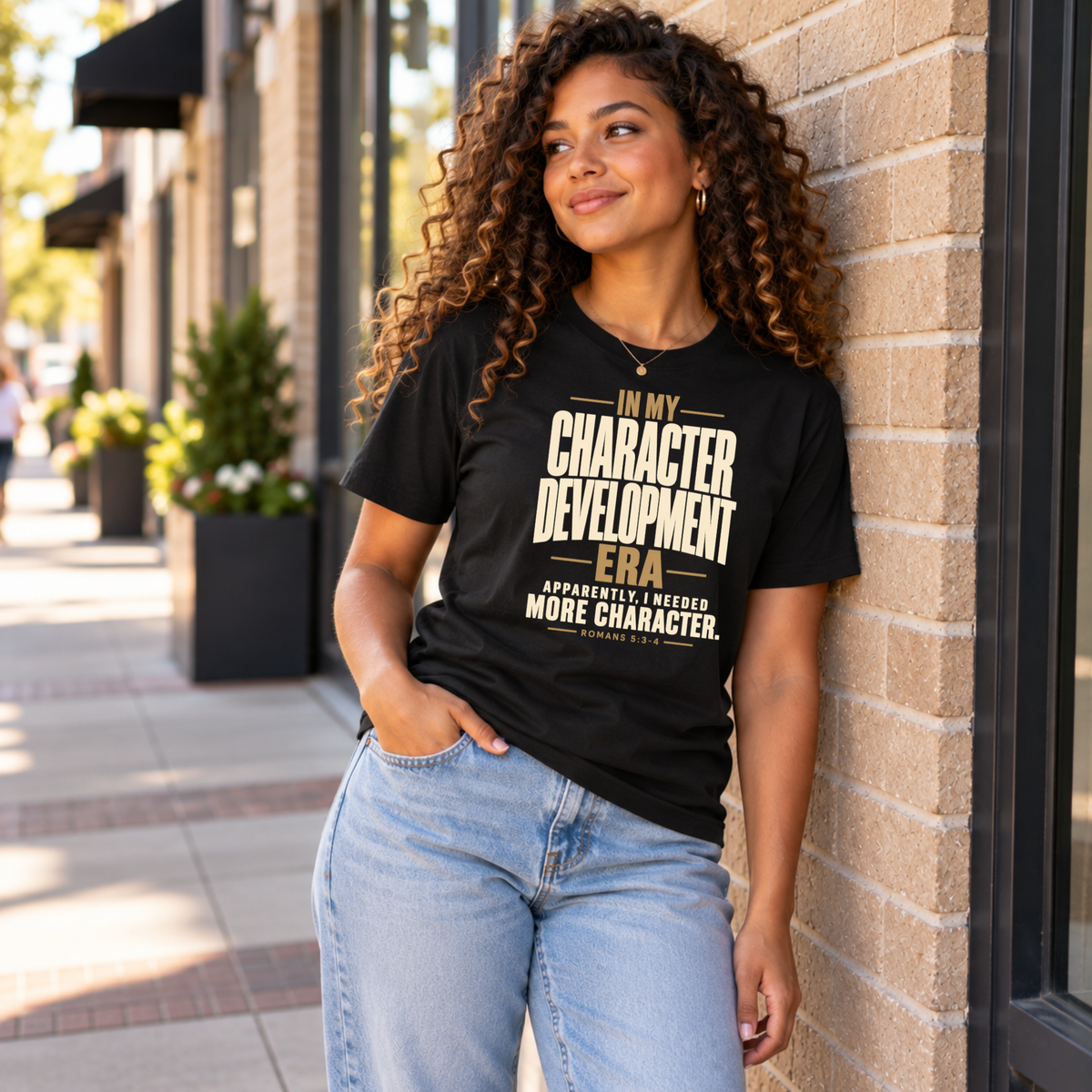 A woman with curly hair leans against a brick building outdoors, wearing a black graphic T-shirt with cream text that reads, 'IN MY CHARACTER DEVELOPMENT ERA Apparently i needed MORE CHARACTER.' She is also wearing light blue jeans.