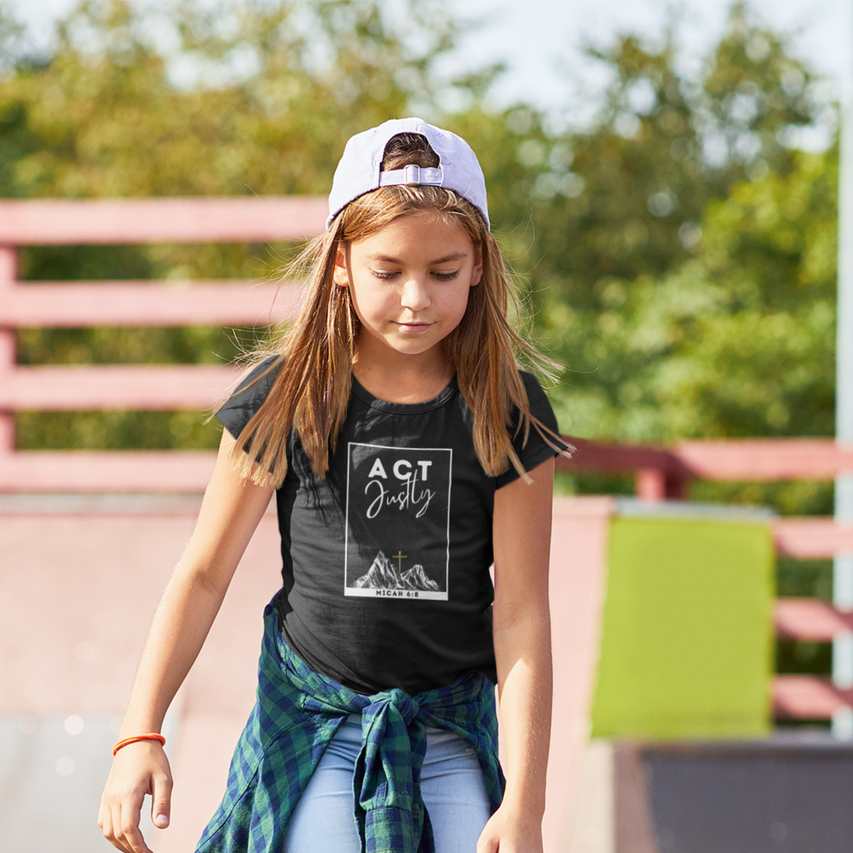 A young girl wearing a black graphic t-shirt with a white 'ACT Justly MINNEVOLS' print, a white cap, and a green plaid shirt tied around her waist, standing outdoors at a skatepark.