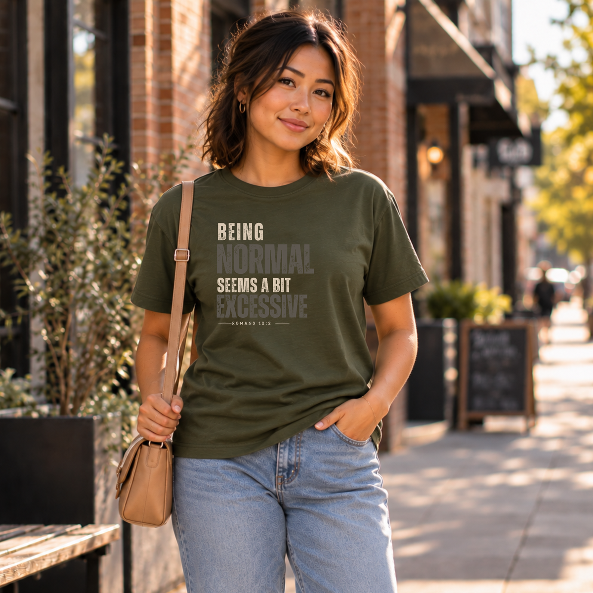 A woman standing on a sidewalk wearing a military green christian graphic shirt that says 'being normal seems a bit excessive' 'romans 12:2'