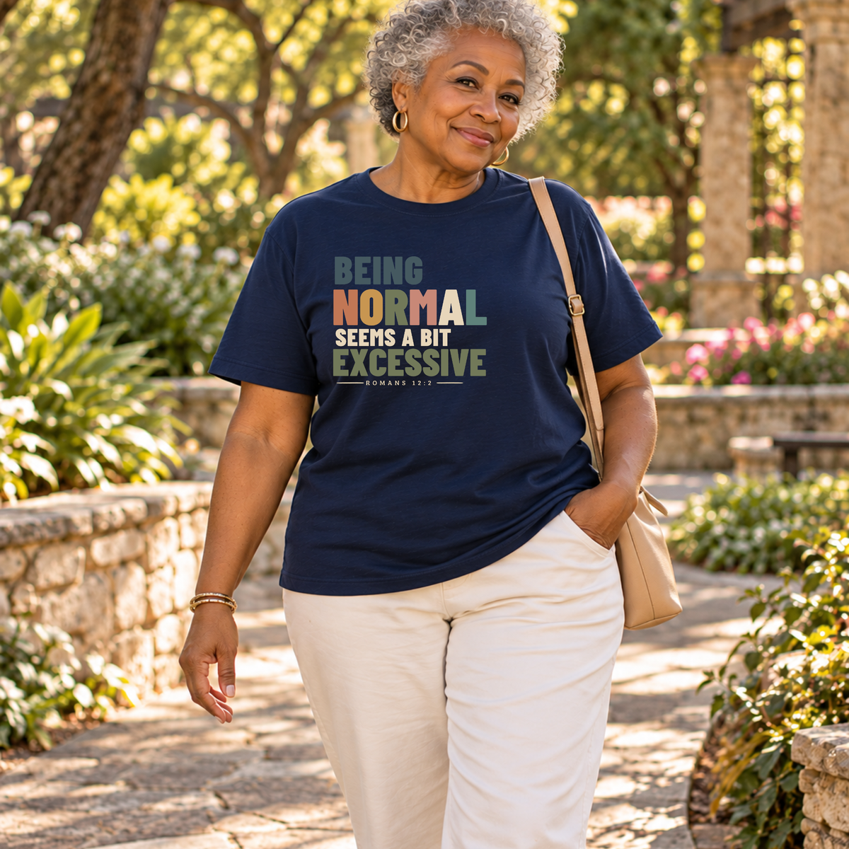 A late 50s woman with curly white hair walking on a sidewalk, wearing a navy blue Christian graphic T-shirt that reads 'BEING NORMAL SEEMS A BIT EXCESSIVE' in multicolor text, paired with light blue jeans.