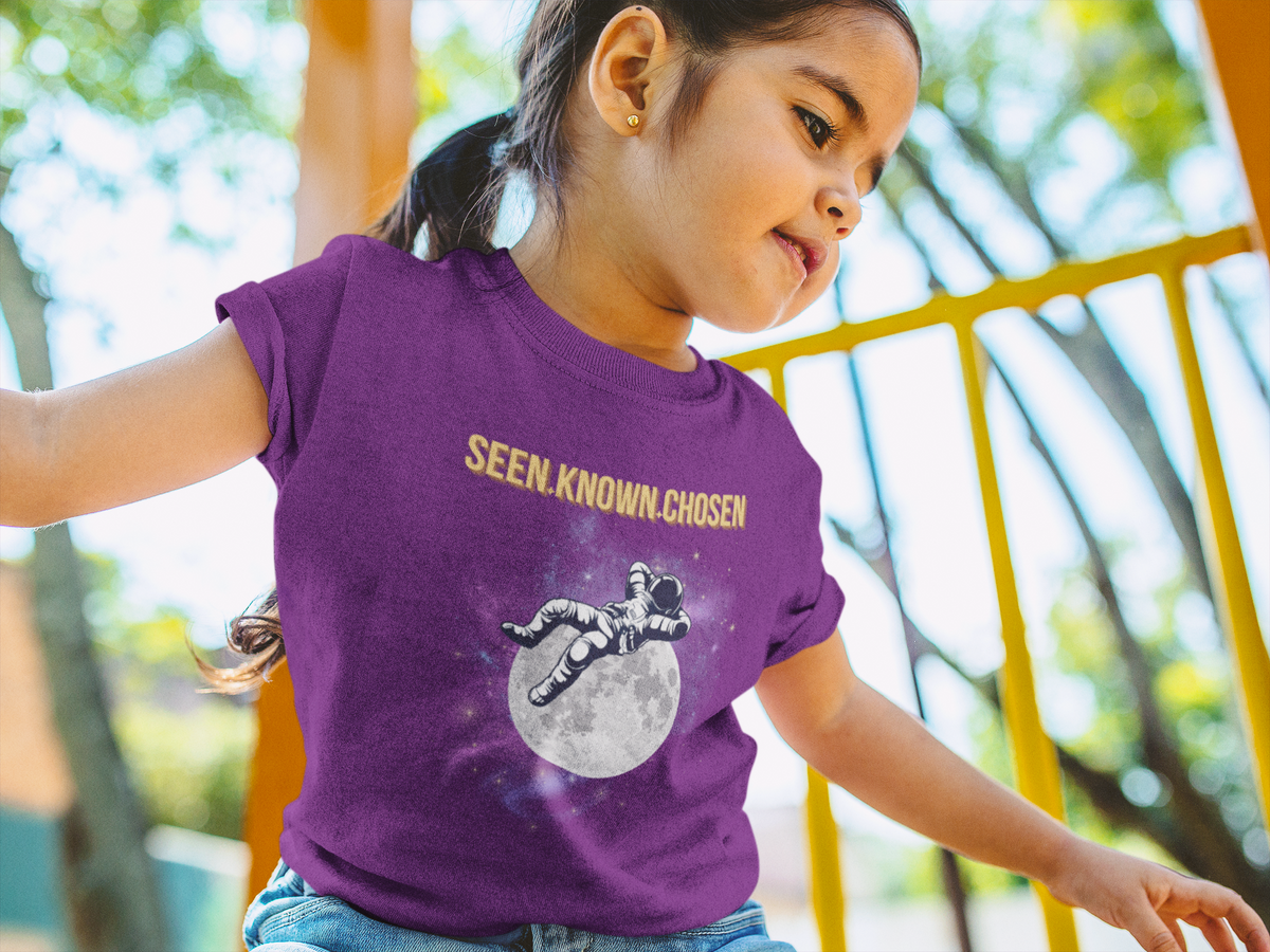 A young girl wearing a purple graphic t-shirt with the text 'SEEN. KNOWN.