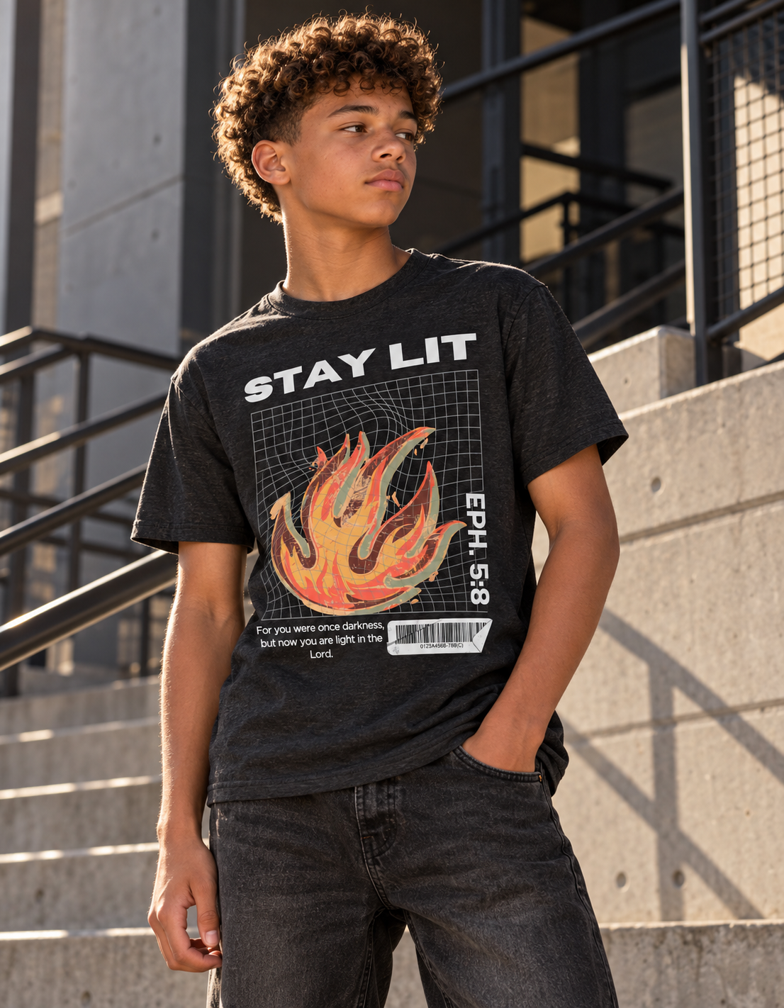 A boy standing around concrete stairs staring off into the distance wearing black jeans and a black Christian shirt that say 'Stay Lit' in big white letters with a retro flame in the center and Eph. 5:8 verticall affixed to the left of the flame and the scripture verse below the flame.