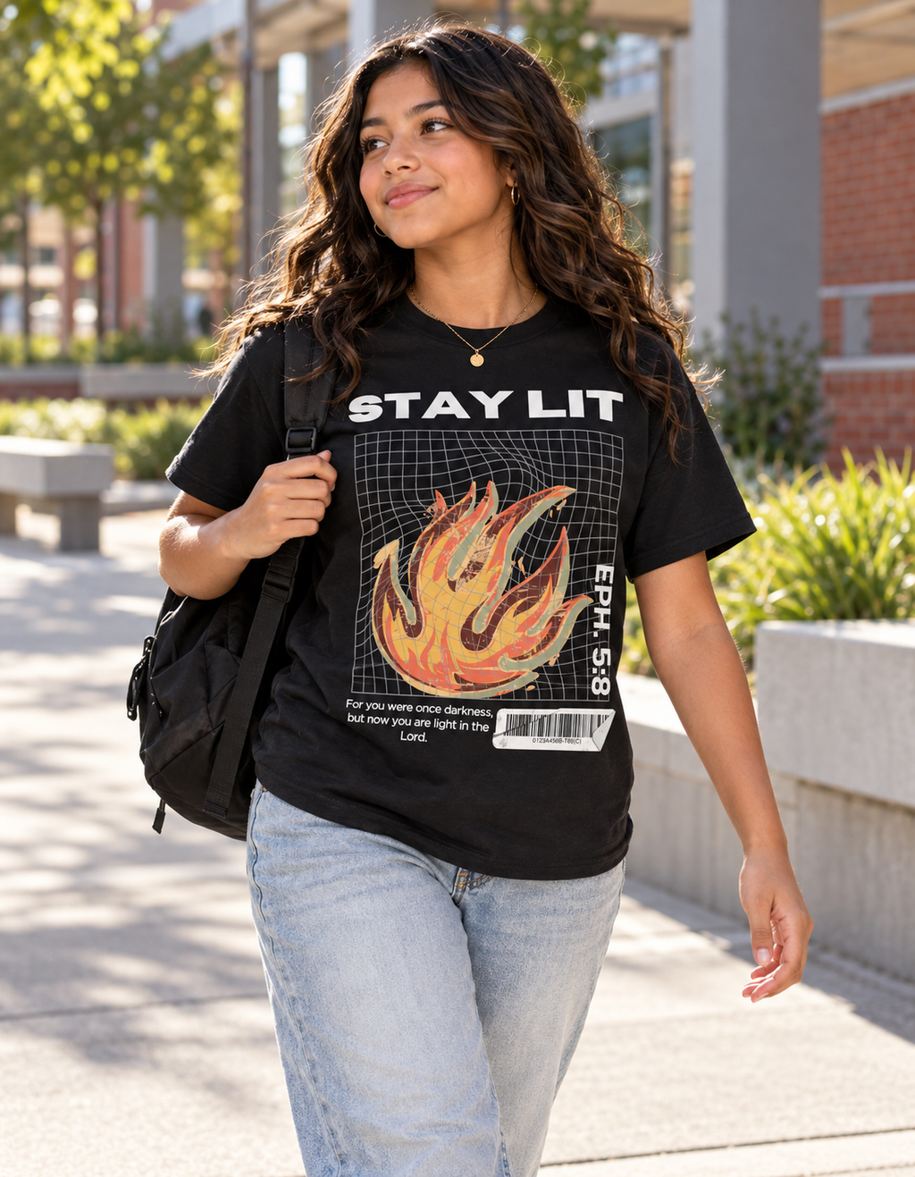 A girl walking outdoors on a sunny day, wearing a black graphic t-shirt with a flame design and the text 'STAY LIT'. She is also wearing light blue jeans and carrying a black backpack.