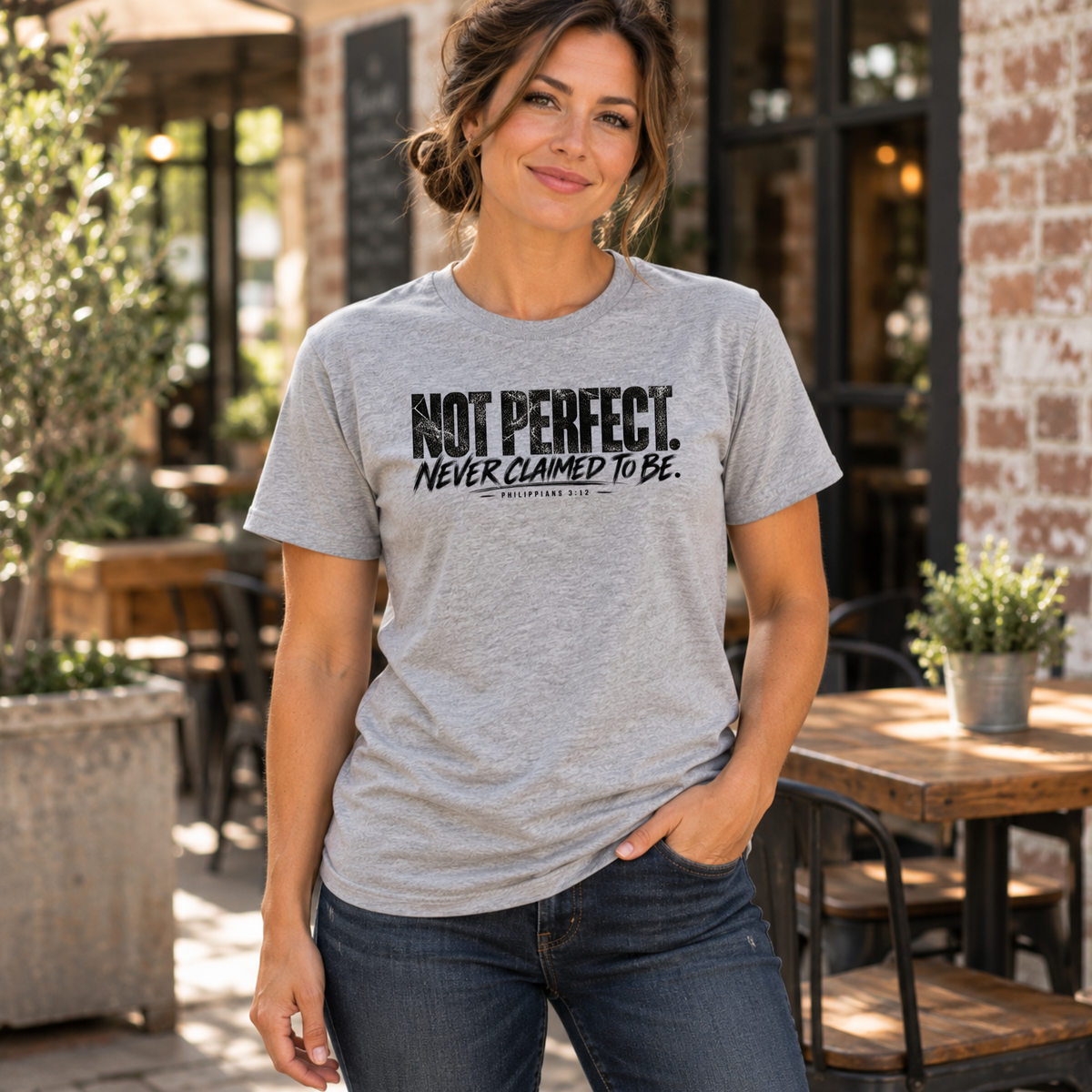 A woman wearing a heather gray graphic t-shirt with the text 'NOT PERFECT. NEVER CLAIMED TO BE.