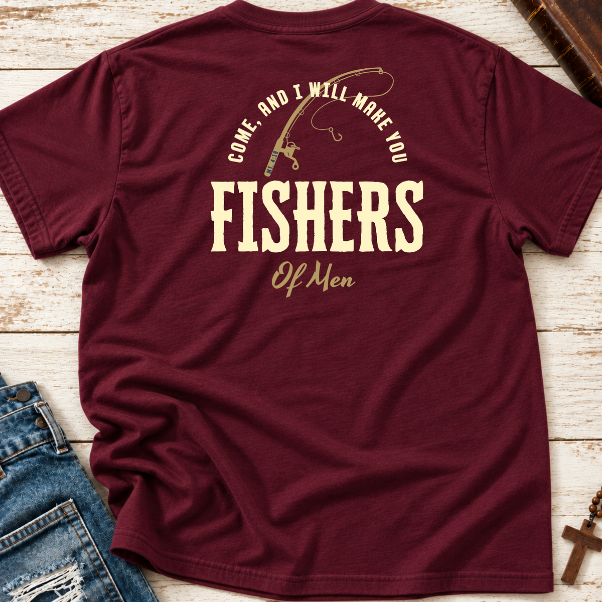 A maroon short-sleeve t-shirt laid flat on a whitewashed wood surface, with blue jeans and a cross visible as styling props. The shirt has a large cream graphic on the back featuring a fishing rod and the text 'COME, AND I WILL MAKE YOU FISHERS OF MEN.