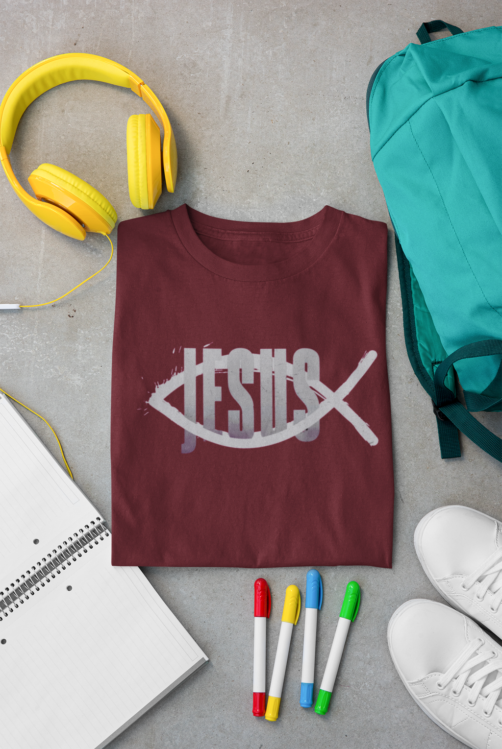 A folded maroon Christian graphic t-shirt by SeedSowerGraphics with a white fish and cross graphic spelling 'JESUS' is arranged in a flat lay on a gray concrete floor, surrounded by yellow headphones, a teal backpack, a notebook, colored markers, and white sneakers.