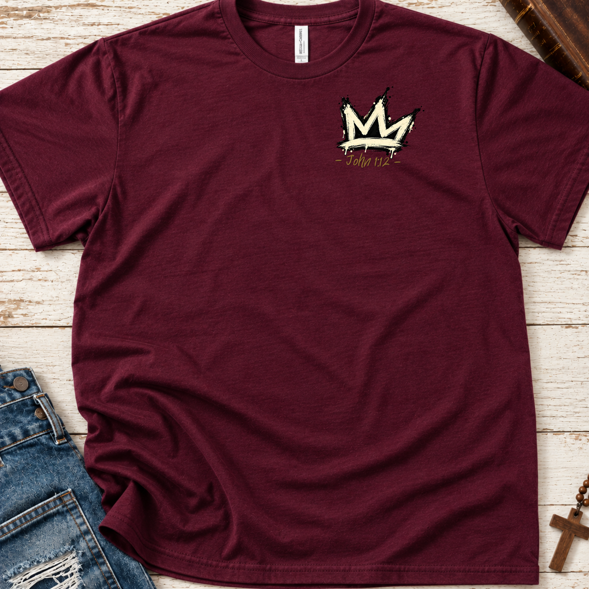 A maroon short-sleeve t-shirt with a gold crown graphic and the text 'John 1:1' on the left chest, styled flat-lay on a white wood background with blue jeans and a cross as props.