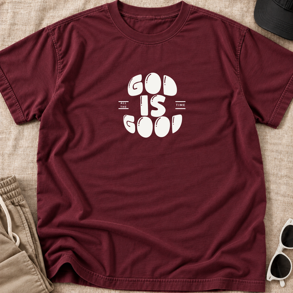 A maroon short-sleeve t-shirt with a white graphic reading 'GOD IS GOOD' laid flat on a beige fabric background, styled with a black cap, khaki pants, and white sunglasses.