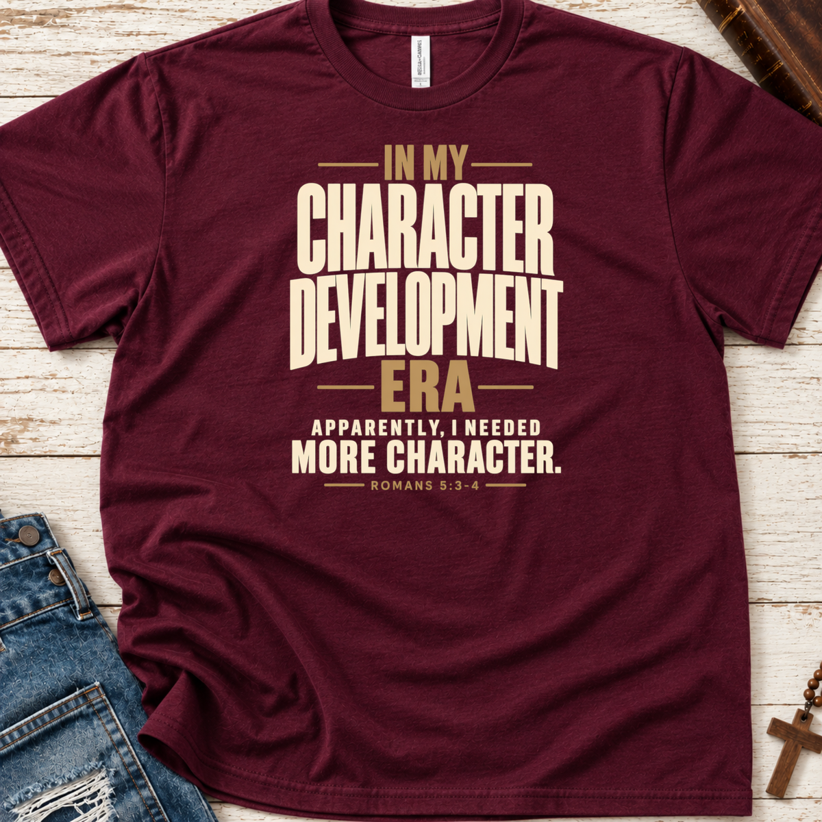 A maroon short-sleeve Christian graphic T-shirt by SeedSowerGraphicsvlaid flat on a whitewashed wood surface, styled with blue jeans, a book, and a cross. The shirt has large cream and tan text reading, 'IN MY CHARACTER DEVELOPMENT ERA APPARENTLY, I NEEDED MORE CHARACTER.