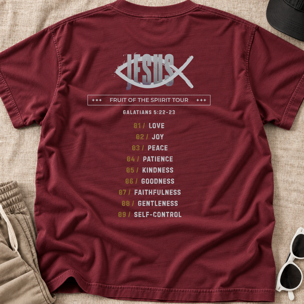 A maroon short-sleeve Christian graphic T-shirt laid flat on a beige fabric surface, with a large back print featuring a fish symbol with 'Jesus' inside, the text 'Fruit of the Spirit Tour,' and a list of nine spiritual virtues with verse Galatians 5:22-23. Partial sunglasses, shorts, and a hat are visible around the shirt as styling props.