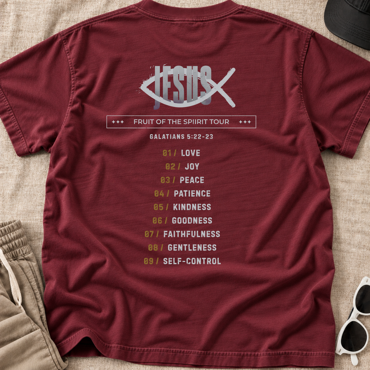 A maroon short-sleeve Christian graphic T-shirt laid flat on a beige fabric surface, with a large back print featuring a fish symbol with 'Jesus' inside, the text 'Fruit of the Spirit Tour,' and a list of nine spiritual virtues with verse Galatians 5:22-23. Partial sunglasses, shorts, and a hat are visible around the shirt as styling props.