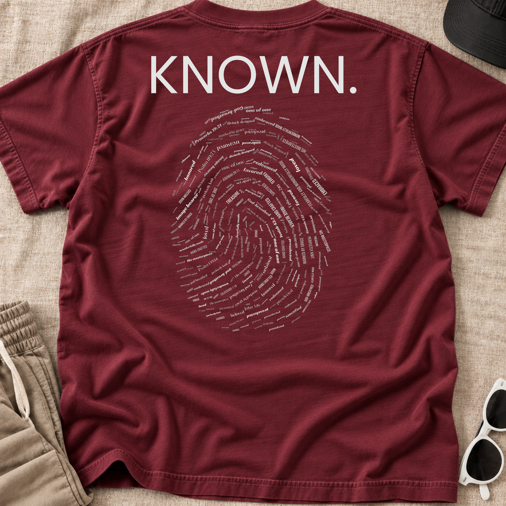 A maroon Christian graphic t-shirt laid flat on a beige fabric background, featuring the word 'KNOWN.' in white block letters above a white fingerprint graphic made of small text.