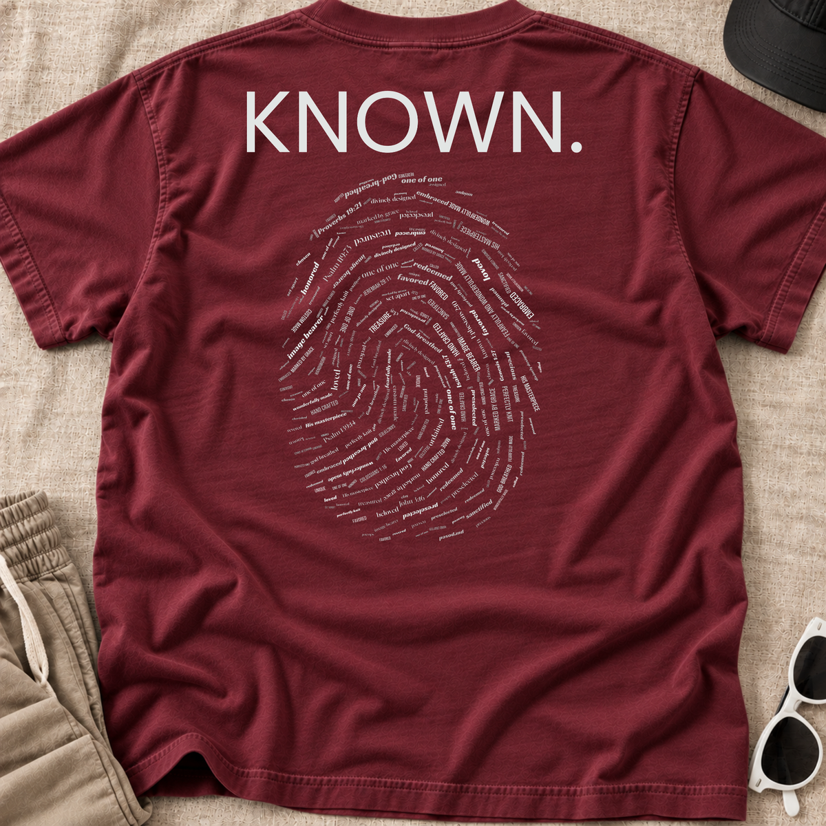 A maroon Christian graphic t-shirt laid flat on a beige fabric background, featuring the word 'KNOWN.' in white block letters above a white fingerprint graphic made of small text.