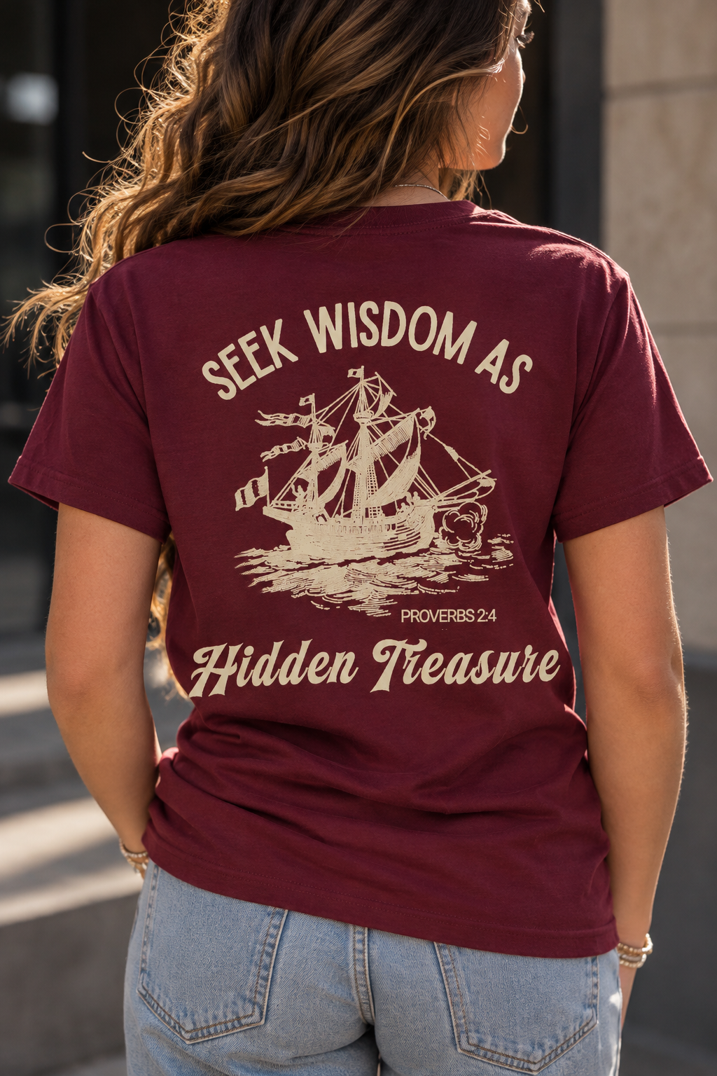 A woman viewed from behind wearing a maroon short-sleeve graphic T-shirt with a cream-colored sailing ship illustration and the text 'SEEK WISDOM AS Hidden Treasure' on the back, paired with light blue jeans.