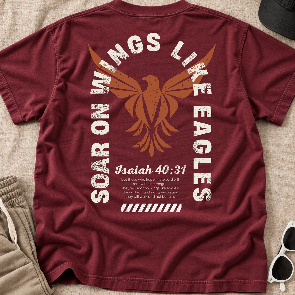 A maroon blue short-sleeve t-shirt laid flat on a light wood surface, showing the back graphic with a large orange eagle, distressed white text reading 'Soar on Wings Like Eagles' and 'Isaiah 40:31', plus smaller scripture text. Khaki bottoms, white sunglasses, and a blackk hat are poart of the props.