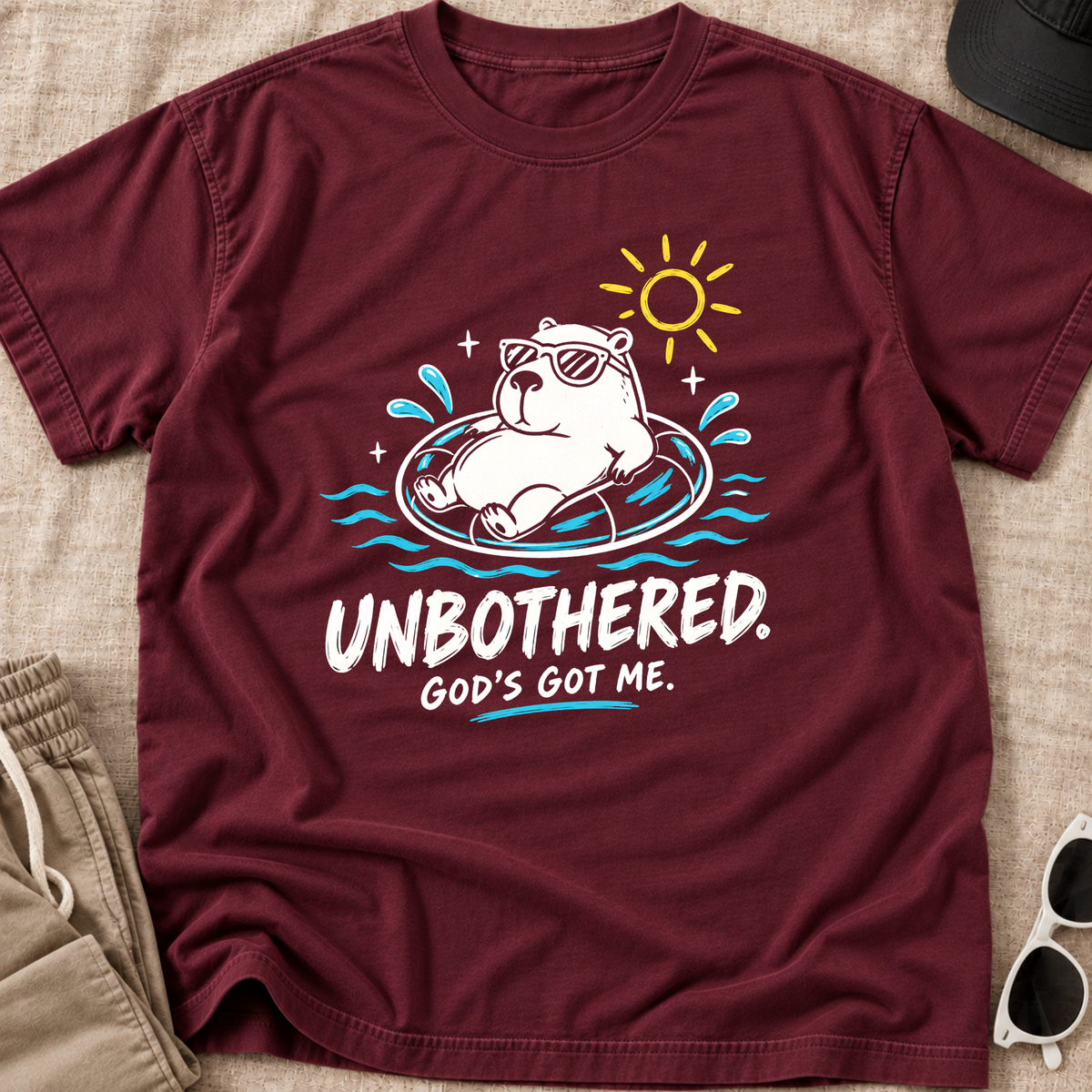 A maroon short-sleeve Christian graphic T-shirt by SeedSowerGraphics laid flat on a beige fabric surface, with a cartoon bear floating in a pool float under a yellow sun and the text 'UNBOOTHERED. GOD'S GOT ME.