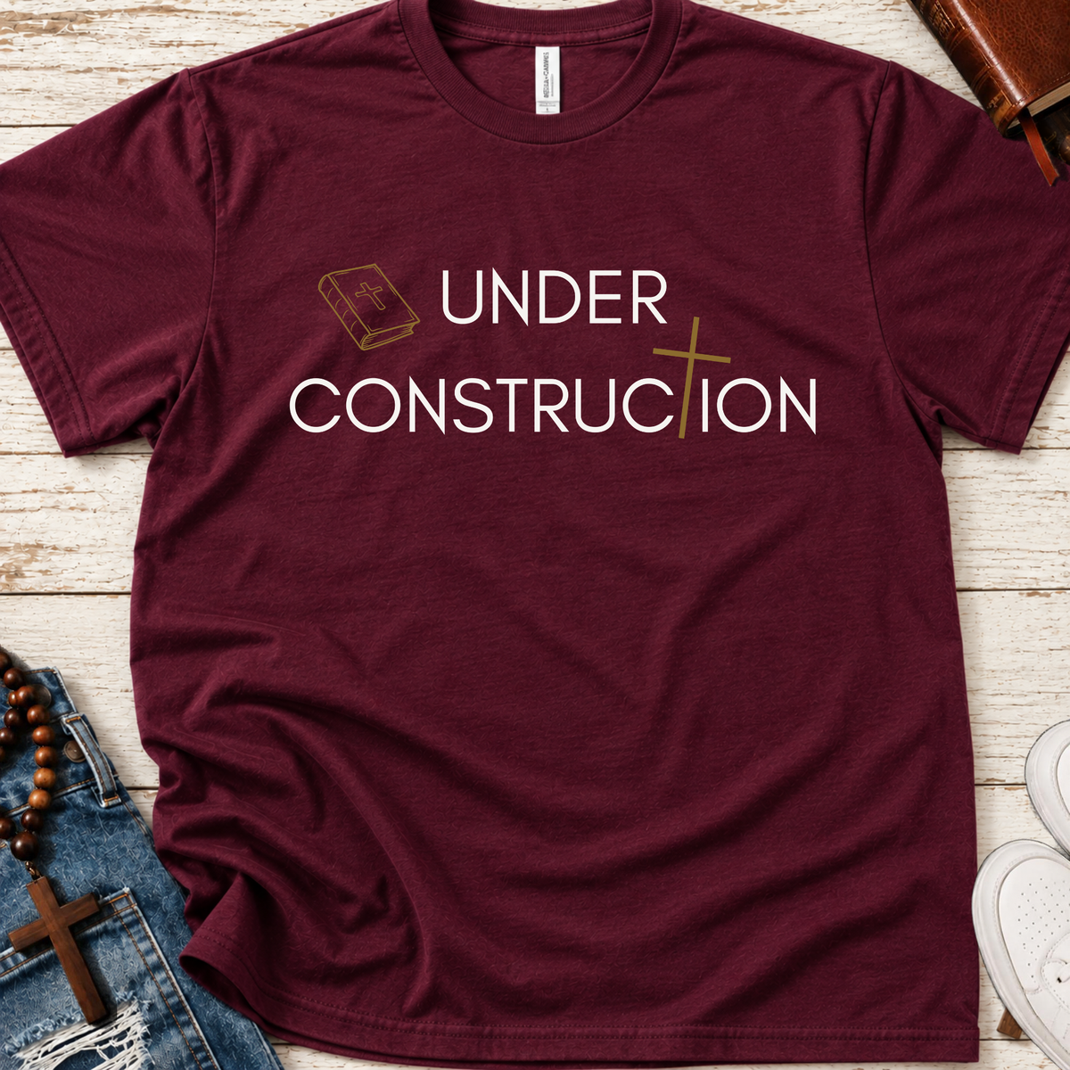 A maroon short-sleeve crewneck t-shirt with a graphic print reading 'UNDER CONSTRUCTION' in white, featuring a small gold book icon on the left and a gold cross on the right. The shirt is displayed in a flat-lay arrangement on a white wood background, surrounded by props including blue jeans, a rosary, a brown leather journal, and white sneakers.