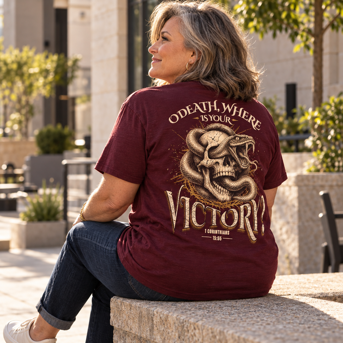 A woman sitting outdoors on a stone bench wearing a maroon graphic T-shirt with a large snake and skull print and the words 'ODATH WHERE IS YOUR VICTORY' on the back.