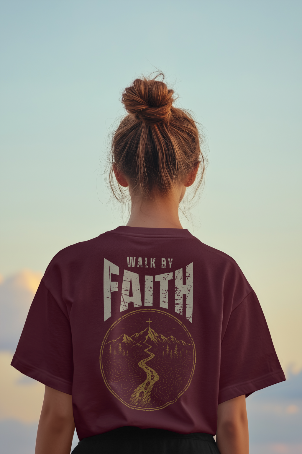 A woman viewed from behind wearing a maroon short-sleeve T-shirt with a large distressed graphic on the back that reads “WALK BY FAITH” above a circular mountain and river illustration. The background is a soft sky gradient in pale blue and beige tones.