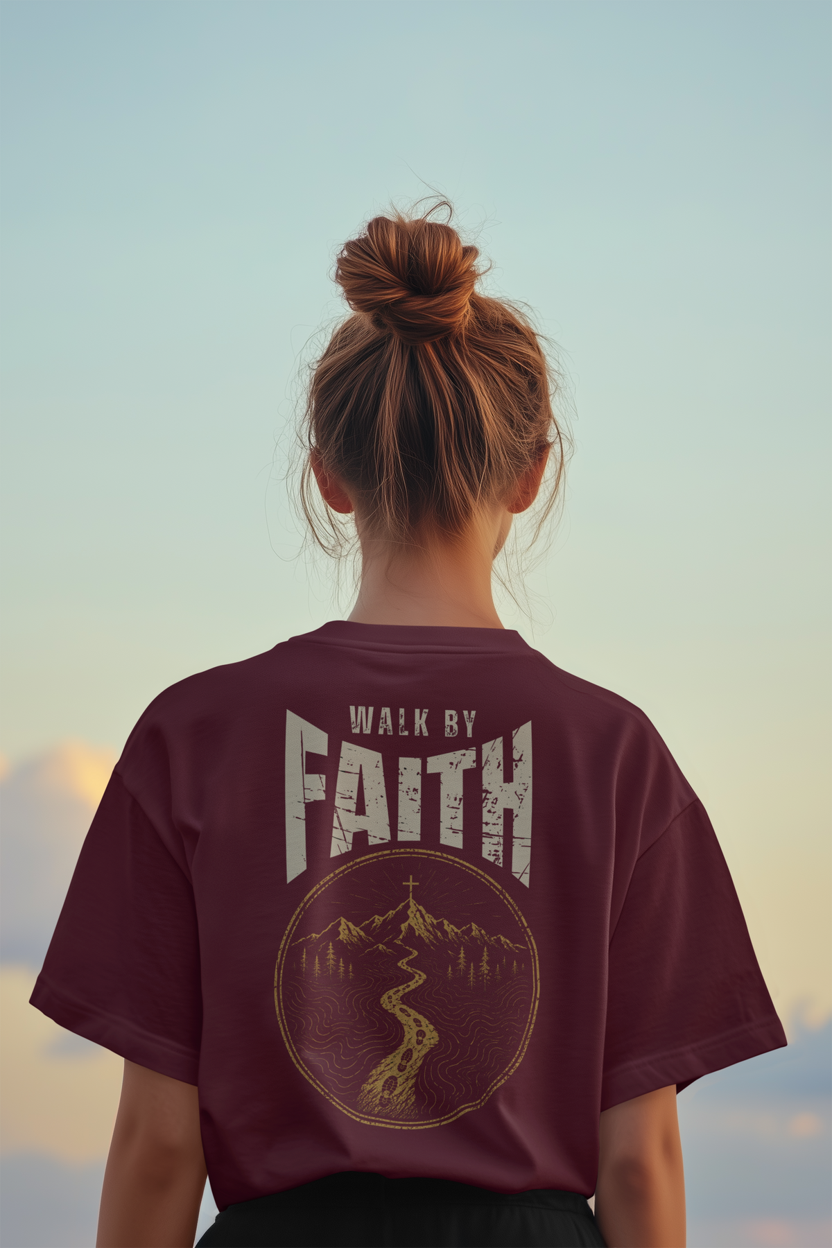A woman viewed from behind wearing a maroon short-sleeve T-shirt with a large distressed graphic on the back that reads “WALK BY FAITH” above a circular mountain and river illustration. The background is a soft sky gradient in pale blue and beige tones.