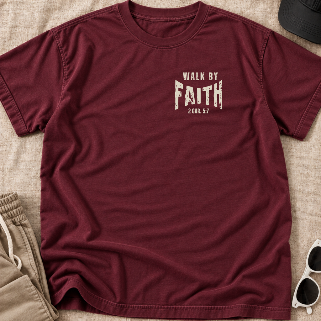 A maroon short-sleeve crewneck t-shirt with a distressed white graphic reading 'WALK BY FAITH 2:008.63' on the chest, laid flat on a beige textured fabric background with partial lifestyle props including sunglasses, a cap, and khaki pants.