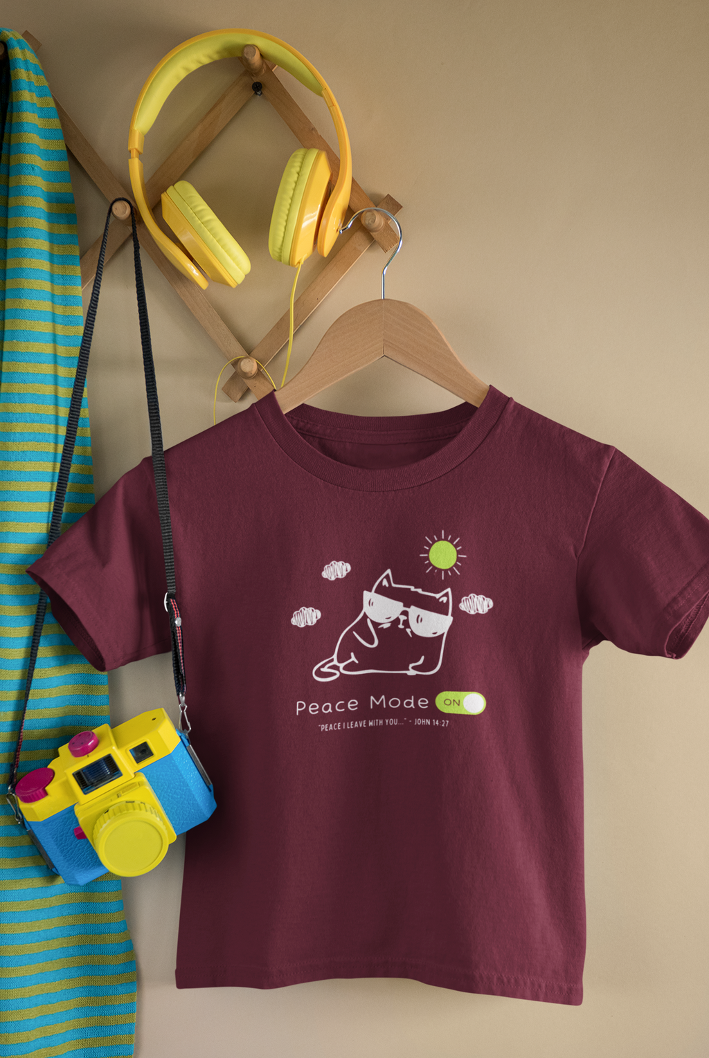 A maroon short-sleeve Christian graphic kids' t-shirt by SeedSowerGraphics on a wooden hanger, featuring a cartoon cat wearing sunglasses with the text 'Peace Mode ON'. The shirt is styled in a room setting with a wooden hanger rack holding yellow headphones, a blue and yellow striped towel, and a toy camera.