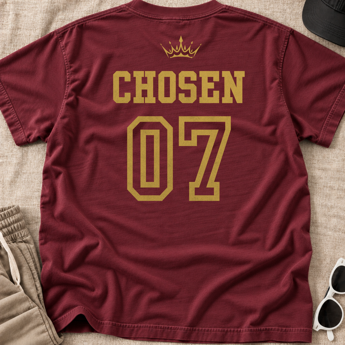 A maroon short-sleeve t-shirt shown from the back, featuring 'CHOSEN' in gold block letters above a large '07' and a small gold crown graphic. The shirt is laid flat on a beige textured fabric background with sunglasses, a black cap, and khaki pants partially visible as styling props.