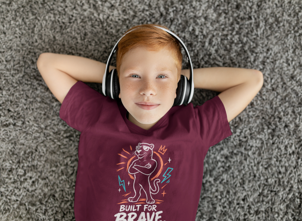 A young boy with red hair lies on a gray shag carpet, wearing a burgundy Christian graphic t-shirt by SeedSowerGraphics with a cartoon tiger and the text 'BUILT FOR BRAVE', along with white over-ear headphones.