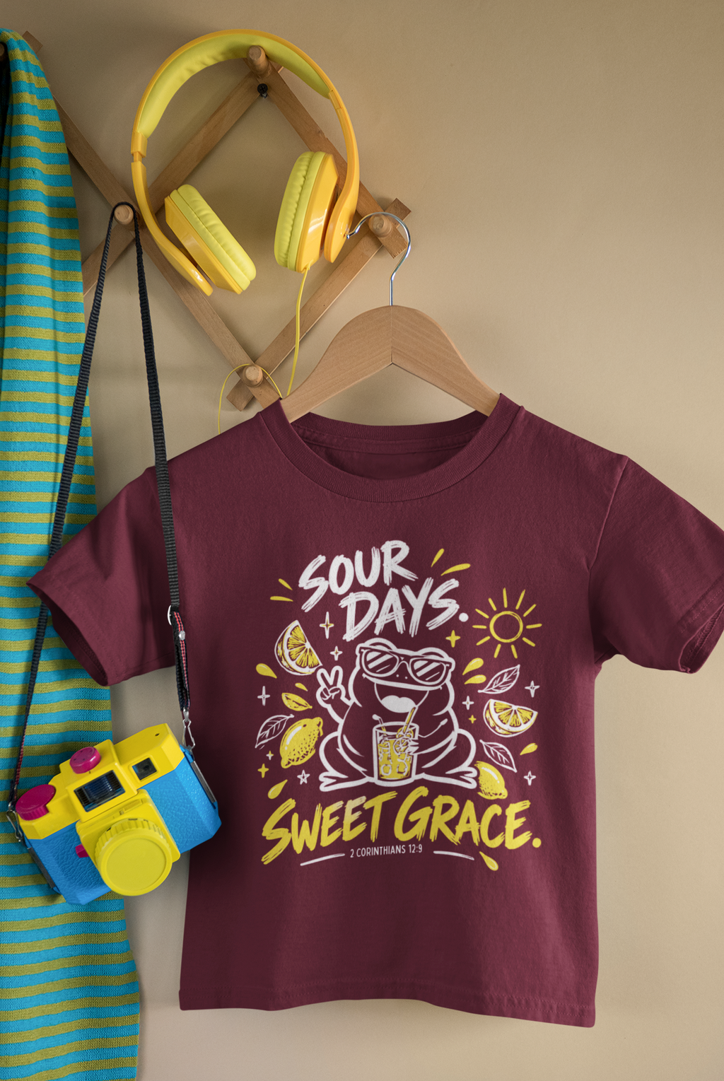 A maroon short-sleeve Christianity graphic T-shirt by SeedSowerGraphics hanging on a wooden hanger against a beige wall. The shirt features a colorful illustration of a frog wearing sunglasses and a drink, with the text 'SOUR DAYS SWEET GRACE.