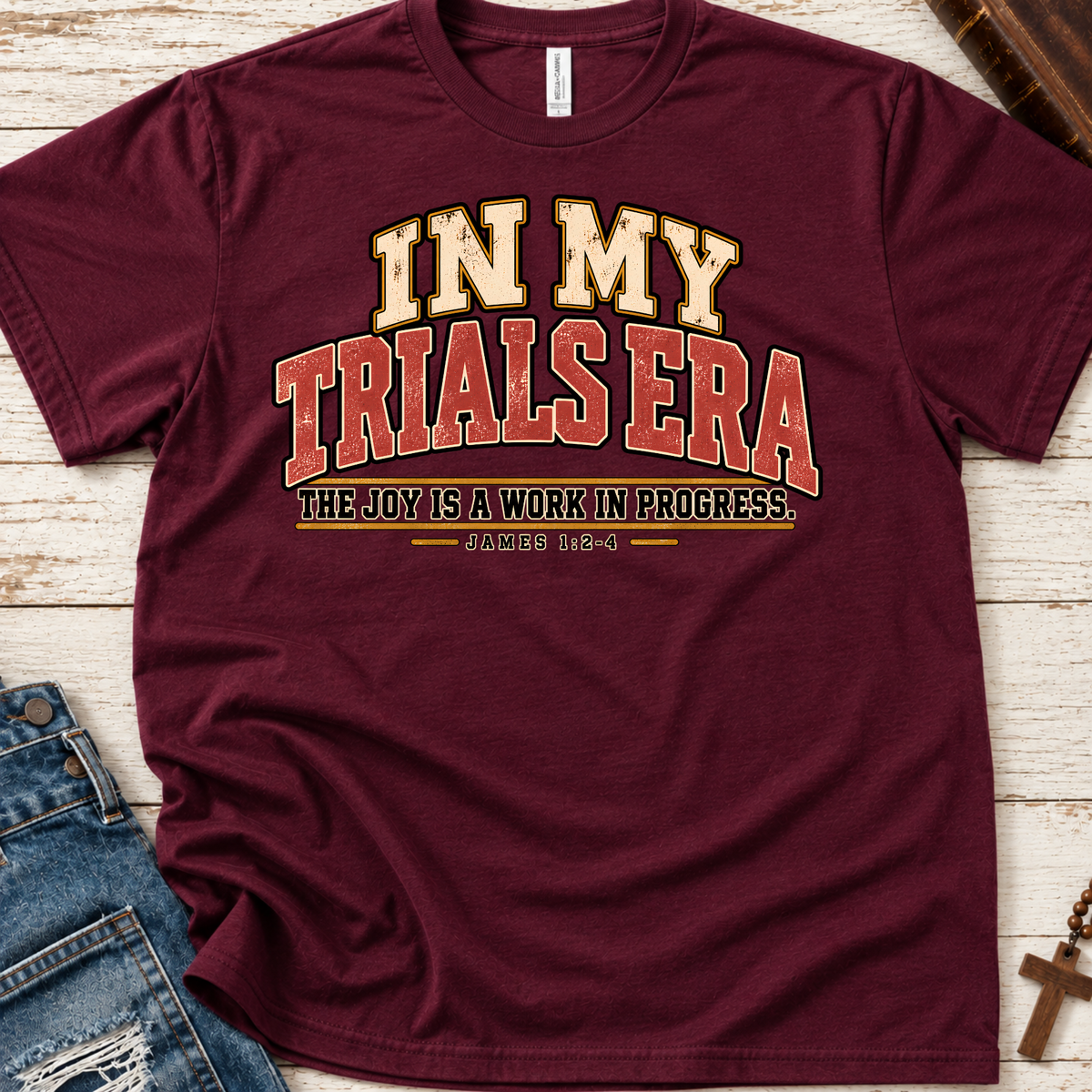 Maroon christian t-shirt by SeedSowerGraphics  laid out amongst props with typography that says "in my trials era' the joy is a work in progress. James 1:2-4 in cream, red, and black lettering