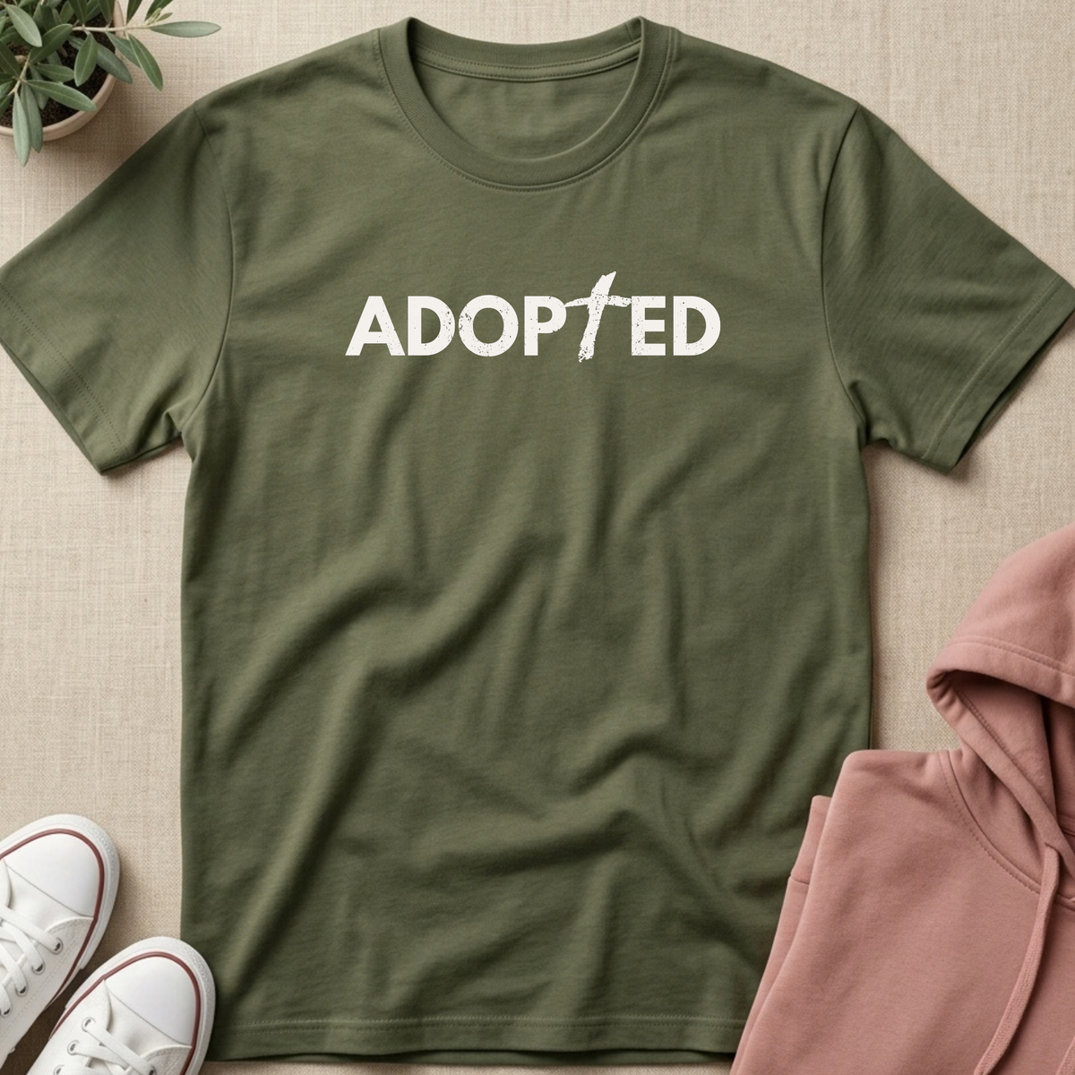 An olive green short-sleeve t-shirt with the word 'ADOPTED' printed in white across the chest, styled in a flat lay with a pink hoodie, white sneakers, and a small potted plant on a beige fabric background.