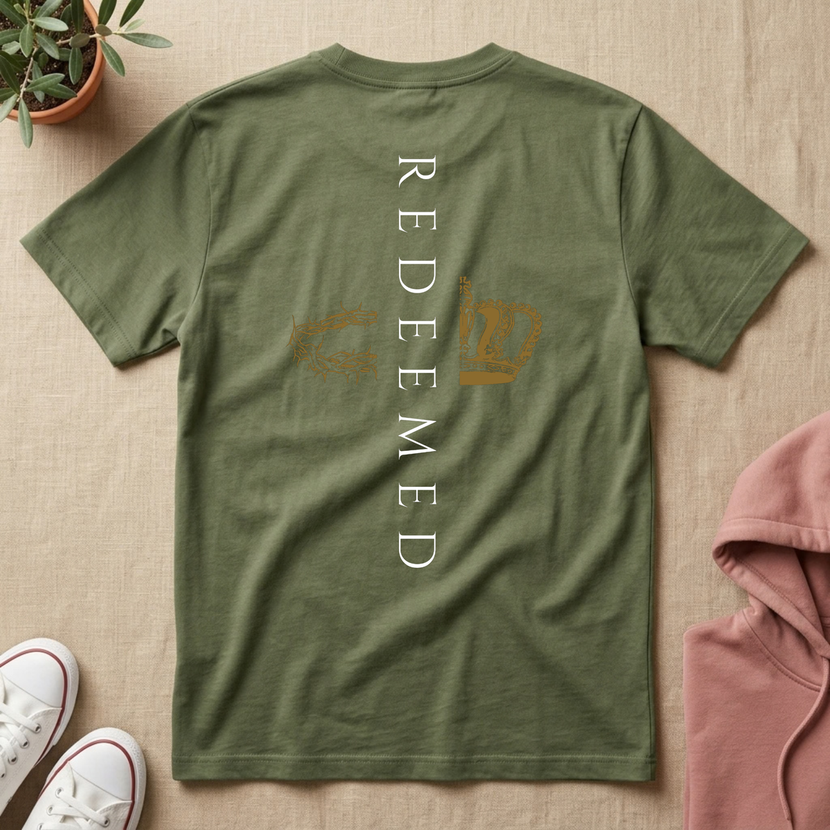 An olive green short-sleeve t-shirt laid flat on a beige fabric background, featuring the word 'REDEEMED' printed vertically in white on the back, with a gold hand graphic and a gold dollar sign. White sneakers, a small potted plant, and a pink hoodie are styled around it.