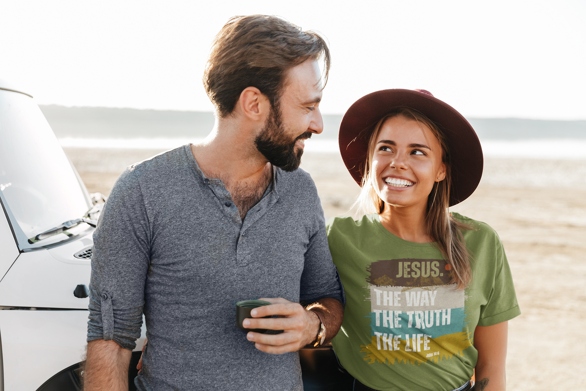 A smiling couple standing outdoors near a white vehicle on a sandy beach. The woman wears a green graphic T-shirt with the text 'JESUS.
