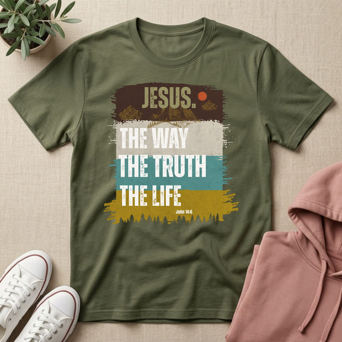 An olive green short-sleeve t-shirt with a graphic print reading 'JESUS. THE WAY THE TRUTH THE LIFE' over a retro multi-colored brushstroke design, presented in a styled flat-lay with a pink hoodie, white sneakers, and a small potted plant on a beige textured background.