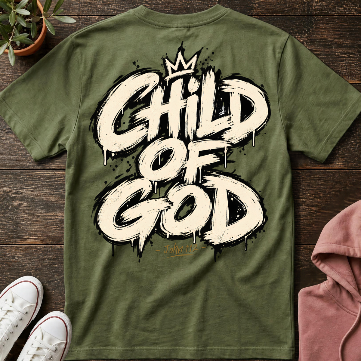 An olive green t-shirt laid flat on a dark wood background, featuring a large white graffiti-style graphic that reads 'CHILD OF GOD' with a small crown at the top. Styled flat-lay props include white sneakers, a pink hoodie, and a potted plant.