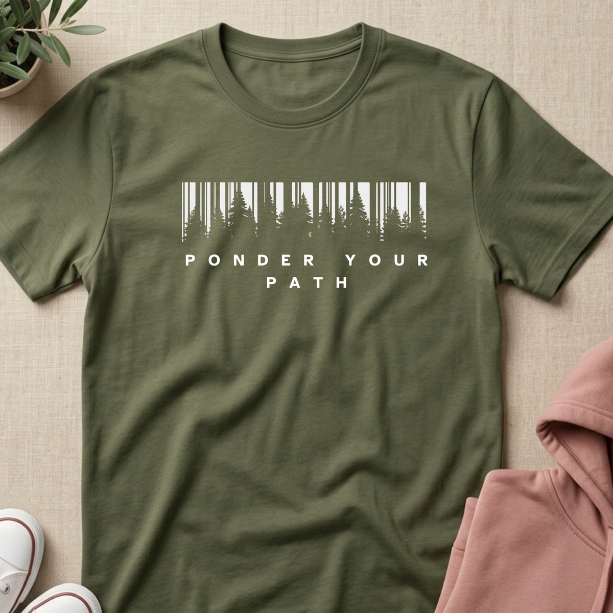 An olive green short-sleeve t-shirt with a white graphic of pine trees and the text 'PONDER YOUR PATH' on the chest, laid flat on a beige textured surface with styled props.