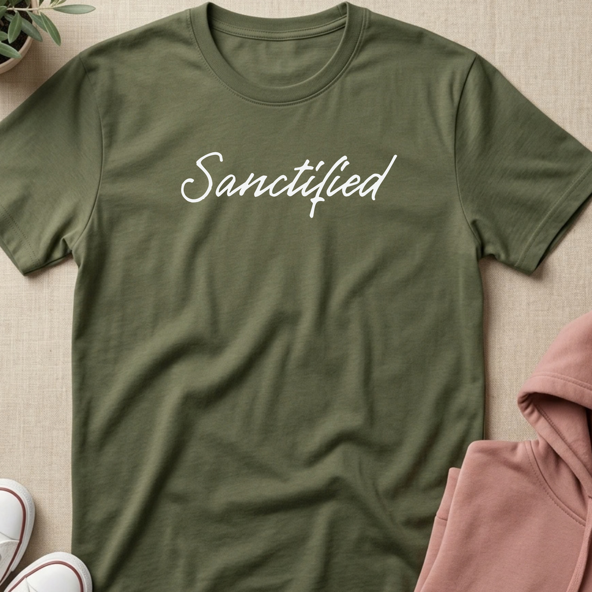 An olive green crewneck t-shirt with the word 'Sanctified' printed in white script across the chest, styled in a flat lay with a pink hoodie, white sneakers, and a small plant on a beige textured background.