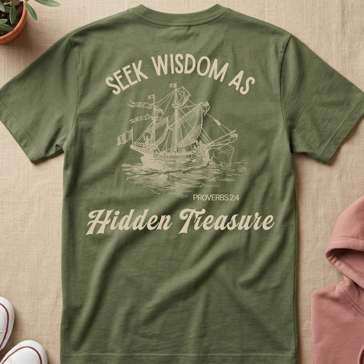 An olive green short-sleeve t-shirt laid flat on a beige textured background, featuring a cream-colored graphic of a sailing ship and the text 'SEEK WISDOM AS Hidden Treasure PROVERBS 2:4'.