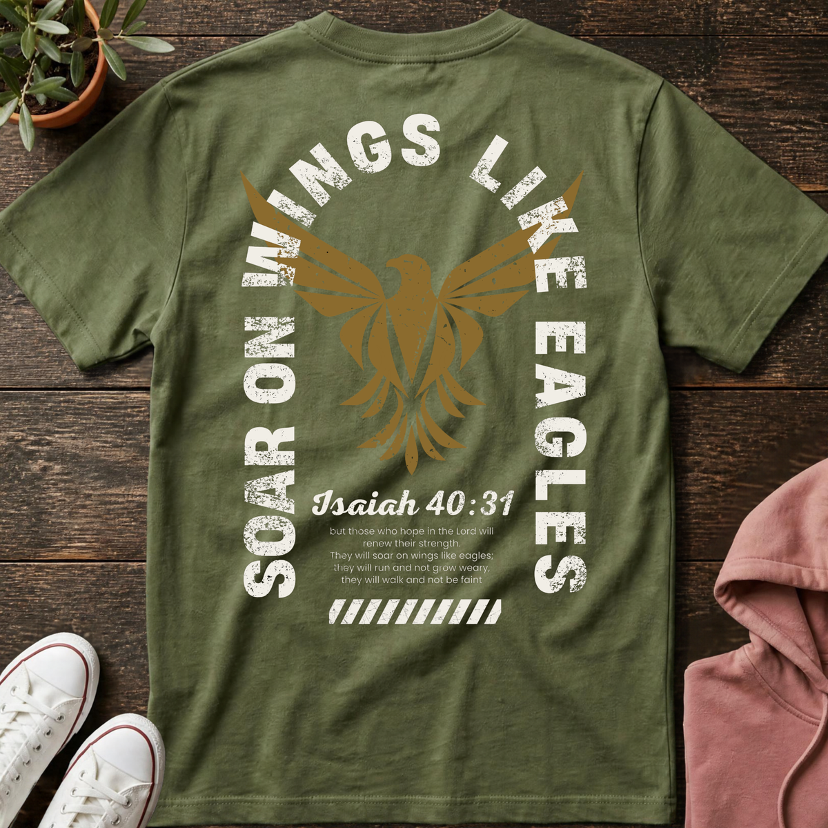 A military green short-sleeve Christian graphic T-shirt by SeedSowerGraphics laid flat on a wood surface, styled with white sneakers, a pink jacket, and a green plant. The shirt features a large distressed gold eagle graphic with white text reading 'SOAR ON WINGS LIKE EAGLES' and 'Isaiah 40:31' printed below.