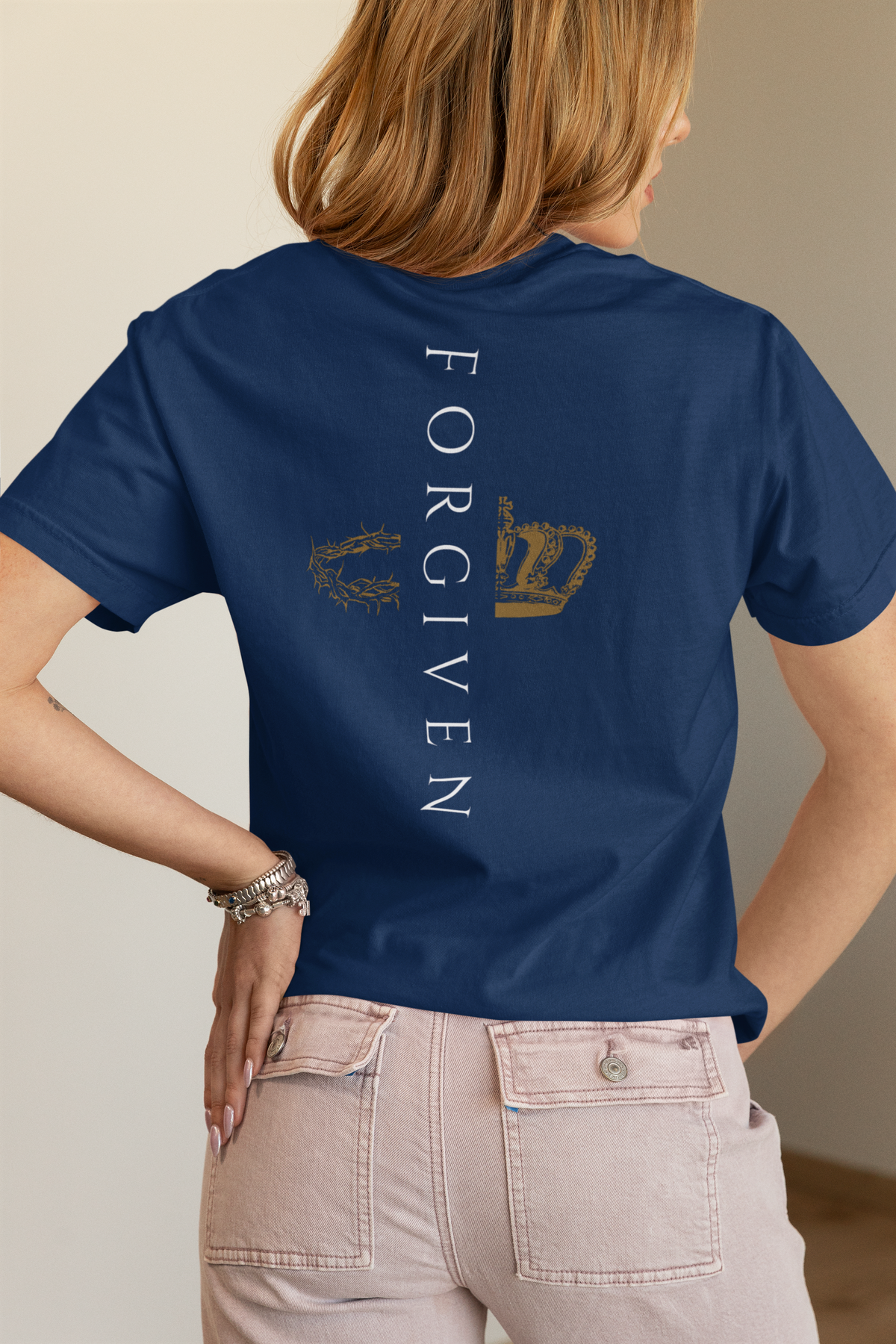 A woman shown from the back wearing a navy blue Christian graphic t-shirt by SeedSowerGraphics with a graphic print on the back. The print features the word 'FORGIVEN' in white vertical text, flanked by a gold laurel wreath on the left and a gold crown on the right.
