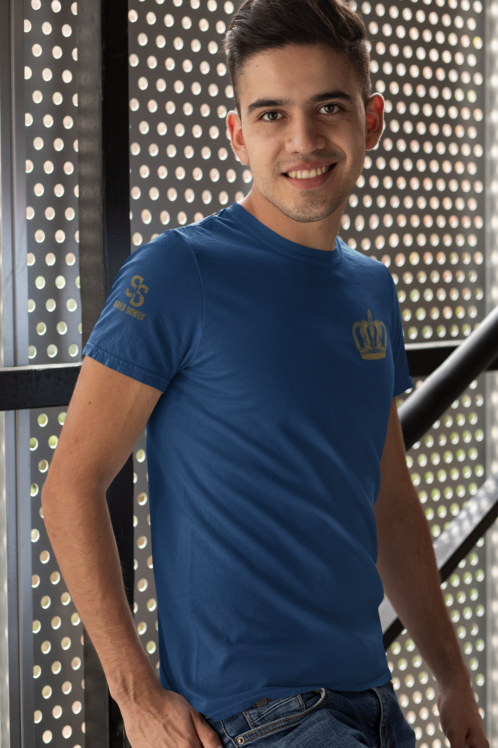 A smiling man wearing a royal blue crewneck Forgiven Christian t-shirt with a gold crown logo on the left chest and a small logo on the right sleeve, standing in front of a perforated metal wall.