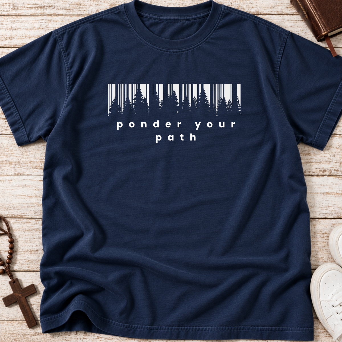 A navy blue crewneck t-shirt with a white graphic print that reads 'ponder your path' below a stylized barcode made of pine trees, laid flat on a light wood background with lifestyle props.