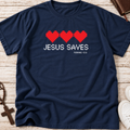 A navy blue short-sleeve t-shirt laid flat on a light wood surface, featuring a graphic with four red pixel hearts and the text 'JESUS SAVES' and 'ROMANS 10:9' in white. A brown cross, rosary, book, and white sneakers are partially visible around the shirt as styled props.