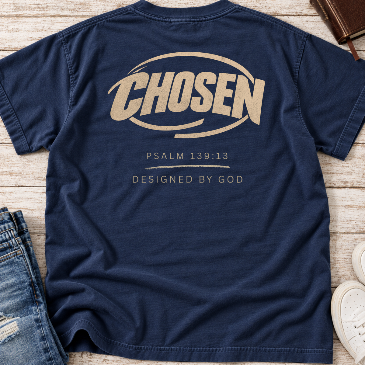 A navy blue t-shirt laid flat on a light wood background, featuring a distressed tan graphic on the back with the word 'CHOSEN' and the text 'PSALM 139:13' and 'DESIGNED BY GOD'.