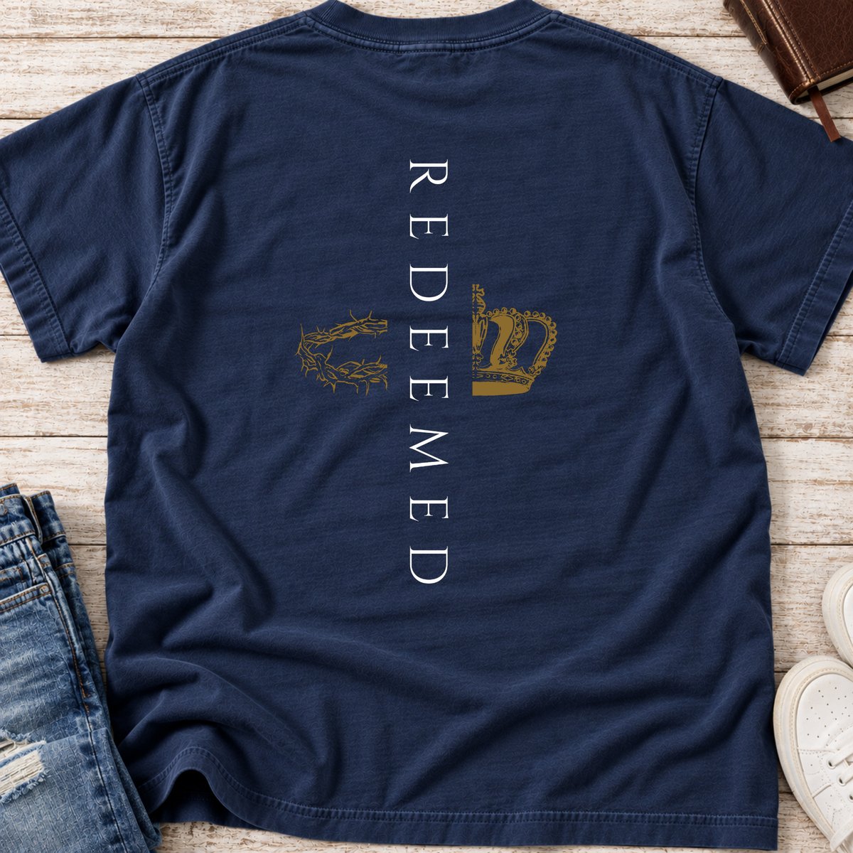 A navy blue short-sleeve Christian t-shirt laid flat on a light wood surface, featuring a vertical white text graphic reading 'REDEEMED' with a gold crown of thorns and a gold crown motif on the chest. Partial jeans, a brown leather item, and white sneakers are visible around the edges as styling props.